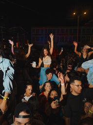 Downtown nighttime outdoor concert crowd, young festivalgoers dancing and cheering with phones raised; woman on shoulders with arms up, wristbands visible.