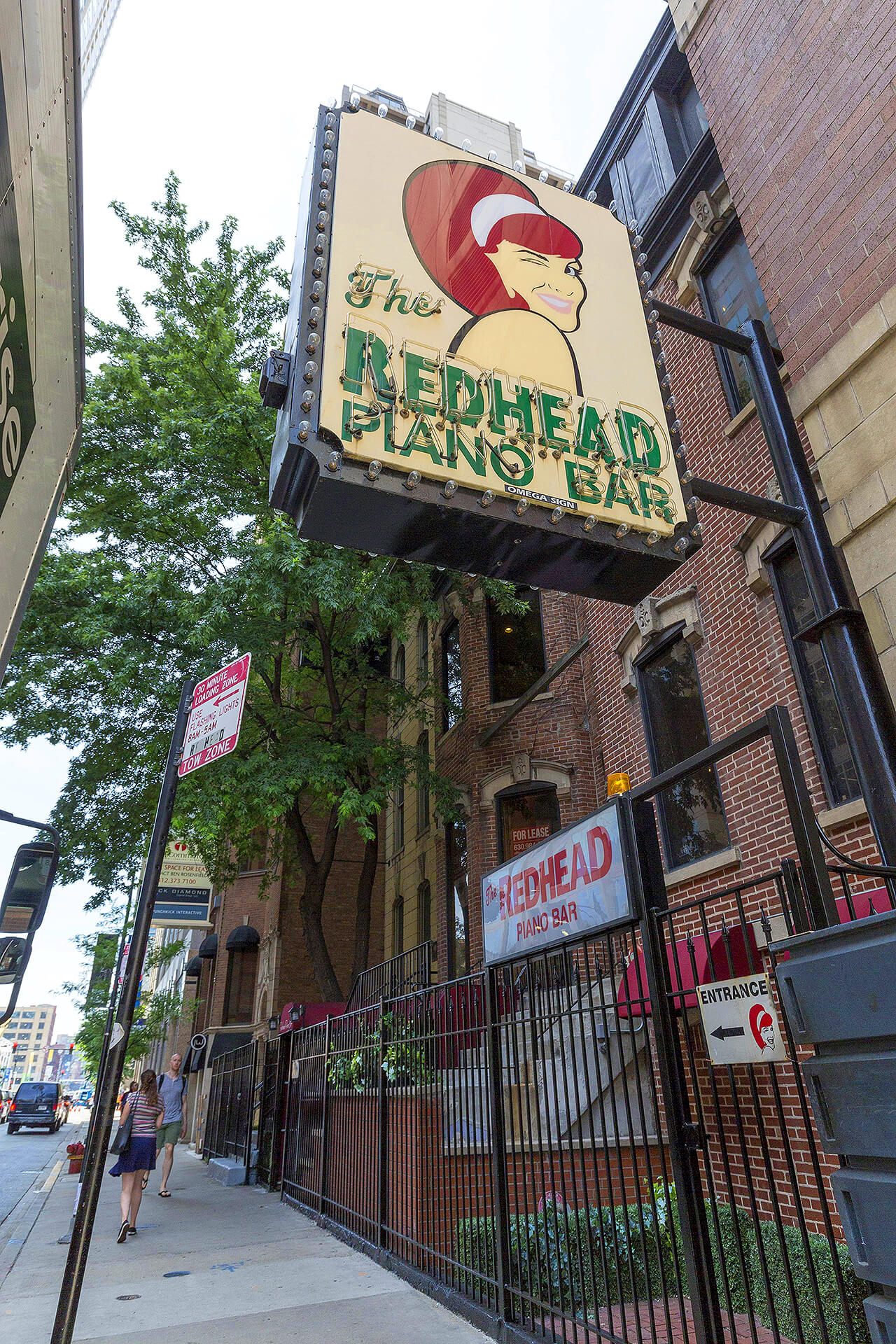 Downtown sidewalk with pedestrians under a large retro marquee featuring a winking red‑haired cartoon and neon lettering for a piano bar mounted on a brick building.