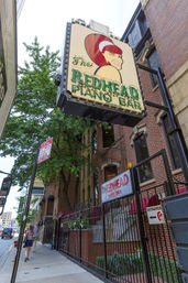 Downtown sidewalk with pedestrians under a large retro marquee featuring a winking red‑haired cartoon and neon lettering for a piano bar mounted on a brick building.