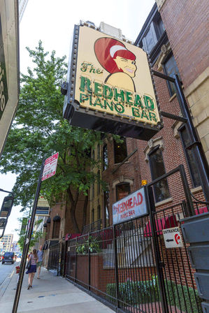Downtown sidewalk with pedestrians under a large retro marquee featuring a winking red‑haired cartoon and neon lettering for a piano bar mounted on a brick building.