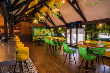 Trendy loft bar interior with vaulted wooden beams, neon-green pendant lights, vibrant green and yellow bar stools and tables, hardwood floor and leafy wall for a cozy urban lounge vibe.