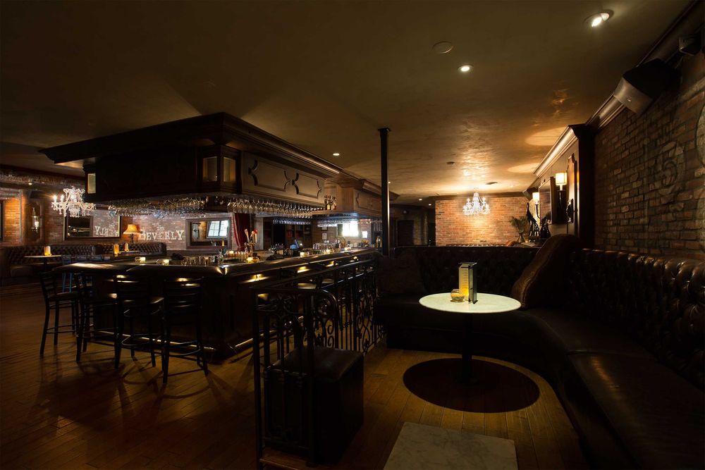 Cozy dimly lit speakeasy-style bar interior with a dark wood U-shaped bar, high stools, tufted leather banquette, small round table with candle, chandeliers and exposed brick walls