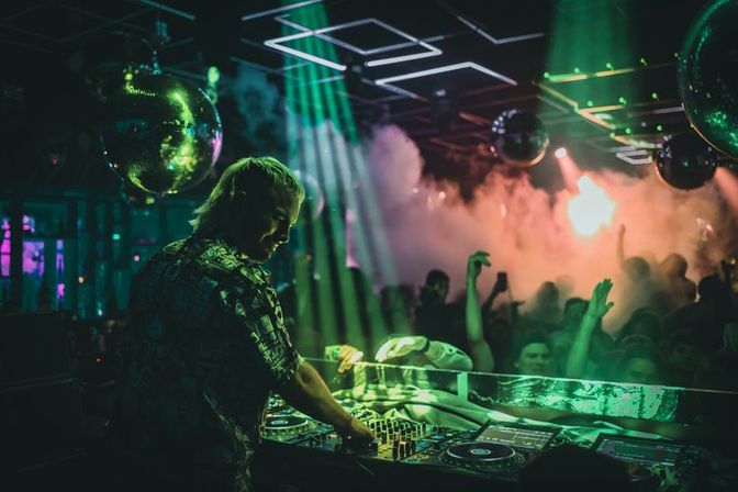 DJ at a nightclub mixing on turntables under green lights and disco balls, smoky dance floor with hands-up crowd and colorful laser beams.