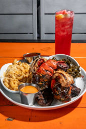 Grilled lobster tail platter with creamy mac and cheese, collard greens and dipping sauces, paired with a bright pink iced cocktail on an orange outdoor table.