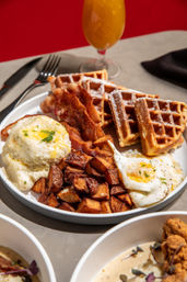 Brunch plate with powdered-sugar waffles, crispy bacon, two sunny-side eggs, seasoned diced potatoes, and a creamy butter-topped biscuit/grits scoop, with a glass of orange juice in the background.