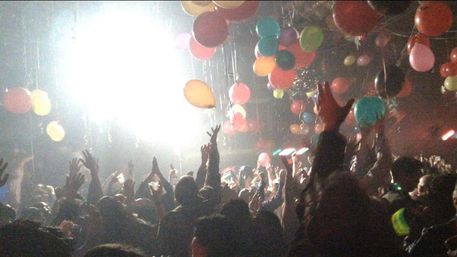 Energetic indoor concert crowd with hands raised under bright stage lights and a colorful balloon drop in a dark club venue, lively party atmosphere.