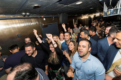 Packed bar nightlife scene with friends dancing, cheering and holding drinks under dim ceiling lights — lively city lounge crowd enjoying a celebration.