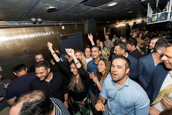 Packed bar nightlife scene with friends dancing, cheering and holding drinks under dim ceiling lights — lively city lounge crowd enjoying a celebration.