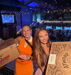 Two smiling friends on an indoor event-venue balcony holding large pizza boxes under string lights, with video screens and a lively night-out crowd below.