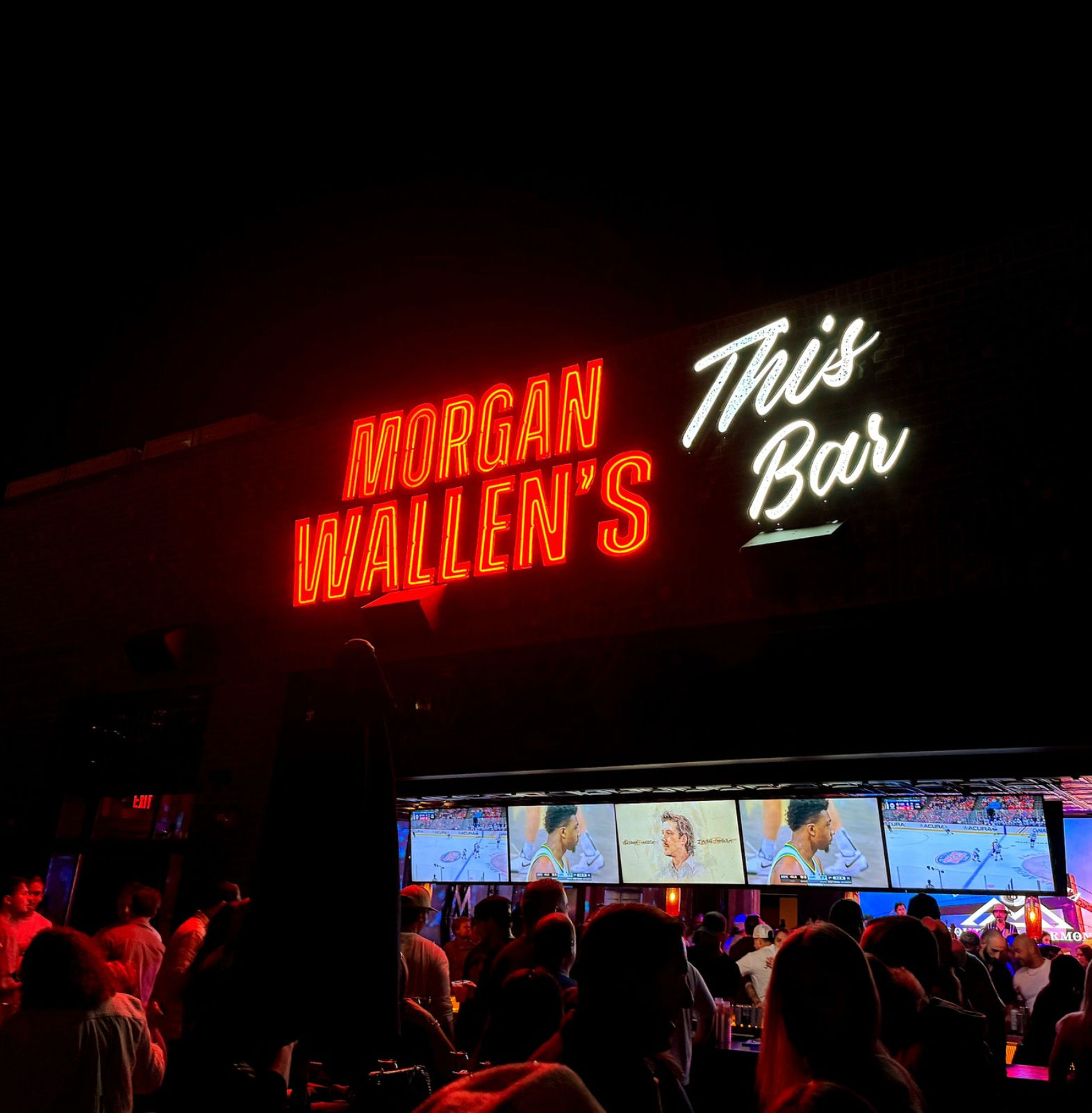 Neon red and white sign showing a country singer’s name and “This Bar” above a lively outdoor sports bar patio at night, crowds watching multiple TVs broadcasting a basketball game.