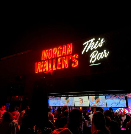 Neon red and white sign showing a country singer’s name and “This Bar” above a lively outdoor sports bar patio at night, crowds watching multiple TVs broadcasting a basketball game.