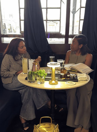 Two women chatting in a cozy urban café booth by a window, sharing drinks, fresh mint, a small lamp and an open book on a round table — casual brunch conversation.