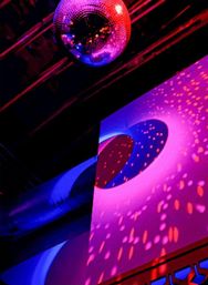 Disco ball reflecting magenta, purple and blue spotlights onto an industrial ceiling duct and curved wall, creating dotted party light patterns in an indoor event space.