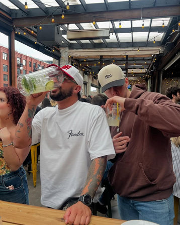 Two friends chugging drinks at a lively downtown rooftop patio with string lights, exposed beams and brick building backdrop.