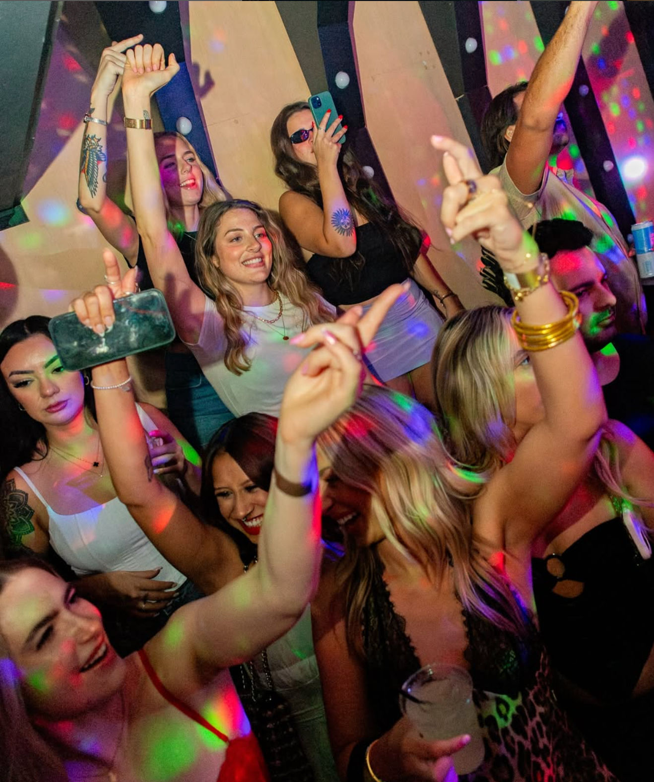 Group of young adults dancing and cheering on a packed nightclub dance floor with colorful disco lights, drinks, and phones capturing the lively nightlife vibe.