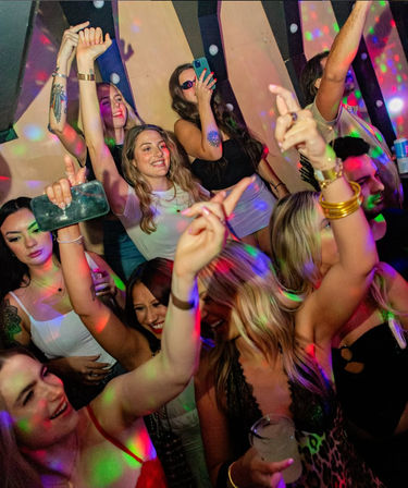 Group of young adults dancing and cheering on a packed nightclub dance floor with colorful disco lights, drinks, and phones capturing the lively nightlife vibe.