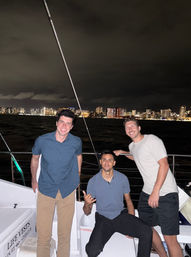 Three friends on a nighttime boat cruise smiling by the rail, one flashing a shaka, with a brightly lit coastal city skyline across the water under a dark, cloudy sky.