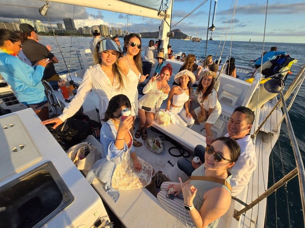 Smiling group of people enjoying a sunset catamaran cruise off Waikiki, Oahu, with the Honolulu skyline and Diamond Head crater on the horizon.