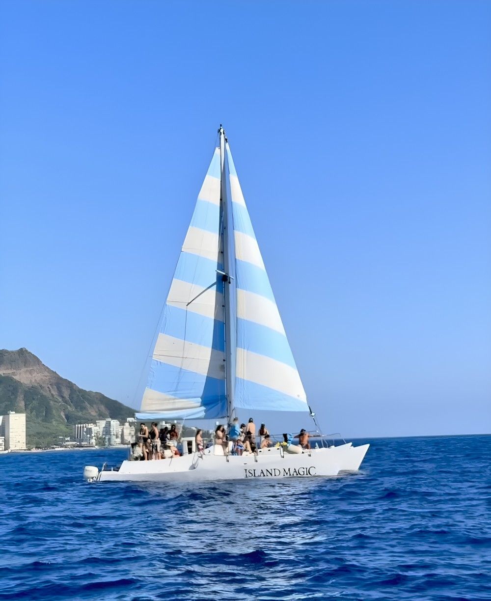 Blue-and-white striped-sail catamaran with passengers sailing on deep-blue Hawaiian waters past a volcanic coastal skyline under a clear sky