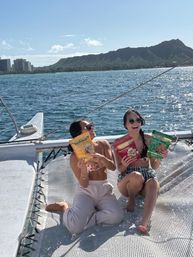 Two women laughing on a catamaran trampoline holding colorful snack bags, sparkling Pacific Ocean and Diamond Head crater on Oahu’s Waikiki coastline in the background on a sunny day