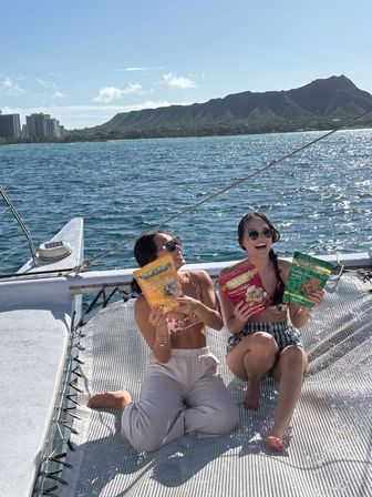 Two women laughing on a catamaran trampoline holding colorful snack bags, sparkling Pacific Ocean and Diamond Head crater on Oahu’s Waikiki coastline in the background on a sunny day