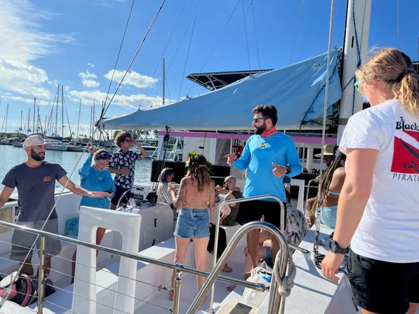 Group of people socializing on a sailboat at a sunny marina, with a furled sail, docked yachts, palm trees and blue sky in the background.