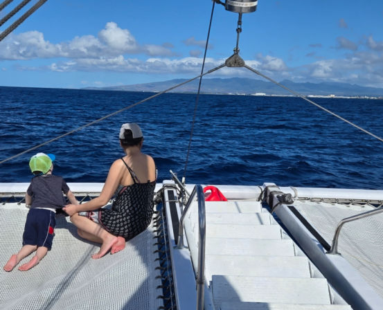 Two people on a catamaran trampoline gazing across deep-blue ocean toward a distant island coastline beneath a sunny, cloud-dotted sky.