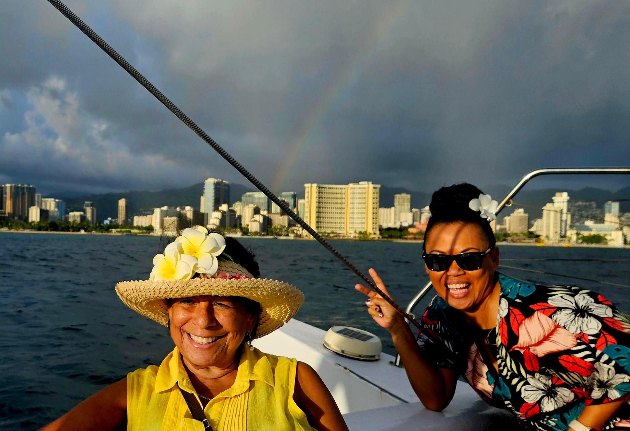 Two smiling women on a boat off a coastal city skyline with a faint rainbow and dark clouds; one in a straw hat adorned with plumeria flowers and a yellow top, the other wearing sunglasses and a colorful floral shirt flashing a peace sign.