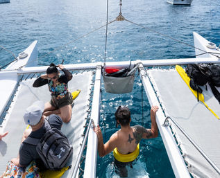 People on a catamaran trampoline preparing to snorkel in clear blue ocean — one person with a snorkel and yellow flotation belt entering the water while others sit on the netted deck with backpacks and a yellow paddle nearby.