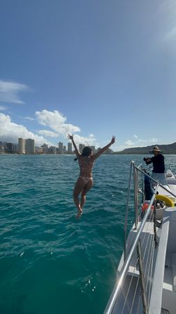 Person in a floral bikini mid-jump off a boat into turquoise Pacific near Waikiki, with the Honolulu skyline and Diamond Head crater under a clear blue sky.