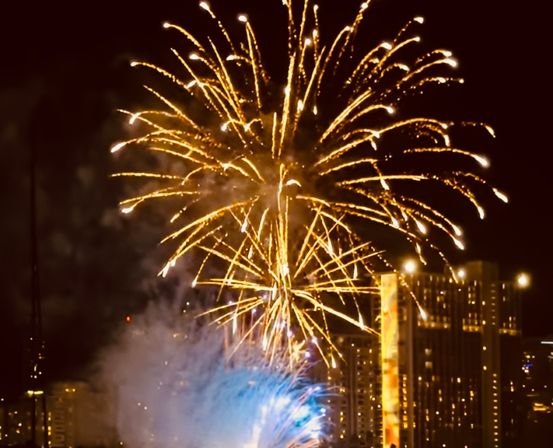 Golden and blue fireworks exploding above a downtown skyline at night, bright streaks and smoke over illuminated high-rise buildings.