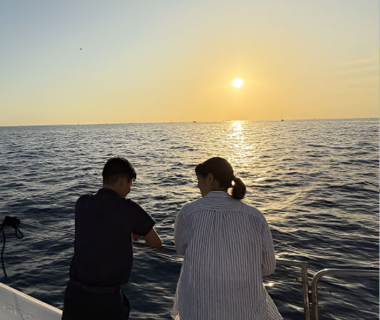 Two people leaning on a boat railing at sunset, watching the sun cast a golden path across the calm open ocean
