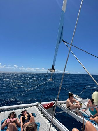 Sunny catamaran sailing on a sparkling deep-blue ocean with friends lounging on the trampoline net and chatting near the bow under a clear blue sky.