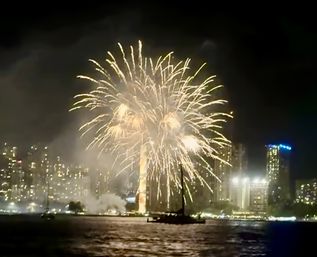Nighttime waterfront fireworks bursting over a city skyline and illuminated high-rise buildings, golden sparks reflecting on the harbor with a silhouetted sailboat in the foreground.