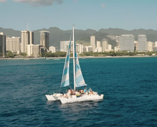 Catamaran with blue-and-white sails and passengers cruising on deep blue Pacific waters off Honolulu, Hawaii, with a coastal skyline and green volcanic ridges in the background.