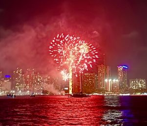Festive red fireworks bursting over an illuminated city skyline at night, colorful explosions and smoke reflecting and rippling across a waterfront harbor.