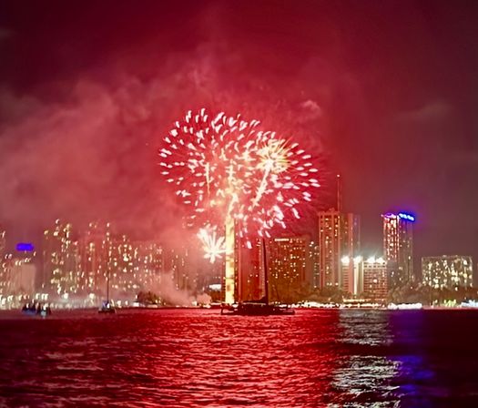 Festive red fireworks bursting over an illuminated city skyline at night, colorful explosions and smoke reflecting and rippling across a waterfront harbor.