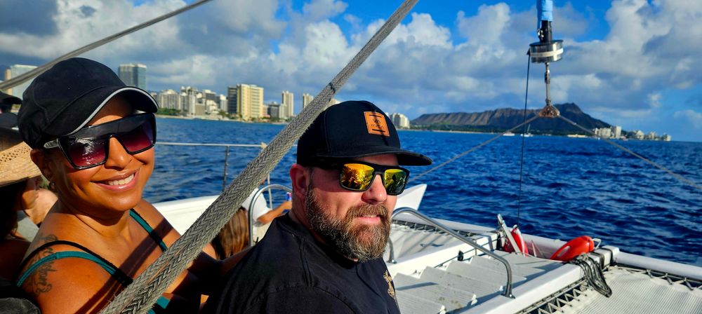 Smiling passengers in sunglasses on a sunlit catamaran with Waikiki skyline and Diamond Head crater on Oahu under bright blue skies