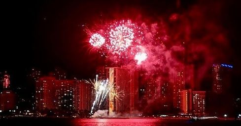 Nighttime urban waterfront with red fireworks bursting over high-rise buildings, vivid reflections on the water and smoky sky — festive city celebration.