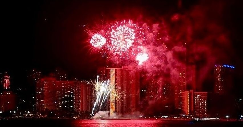 Nighttime urban waterfront with red fireworks bursting over high-rise buildings, vivid reflections on the water and smoky sky — festive city celebration.
