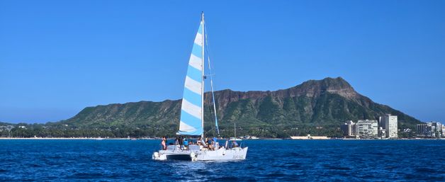 White catamaran with blue-striped sail cruising deep blue Pacific waters off Waikiki, Honolulu, with Diamond Head crater and beachfront skyline under a clear sunny sky