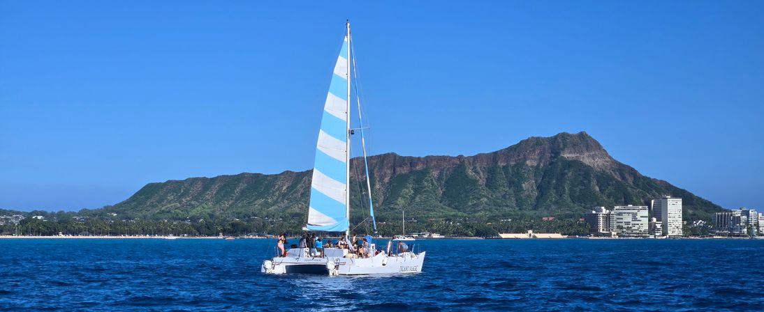 White catamaran with blue-striped sail cruising deep blue Pacific waters off Waikiki, Honolulu, with Diamond Head crater and beachfront skyline under a clear sunny sky