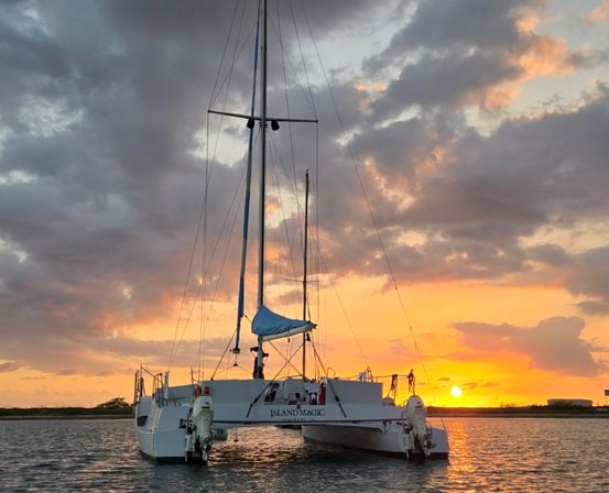 White catamaran anchored in calm coastal water at a vibrant orange-pink sunset with dramatic clouds.