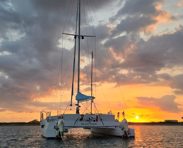 White catamaran anchored in calm coastal water at a vibrant orange-pink sunset with dramatic clouds.