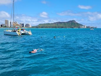 Snorkelers and a white catamaran on vibrant turquoise water off Waikiki, with Honolulu high-rises and Diamond Head crater under a sunny blue sky.