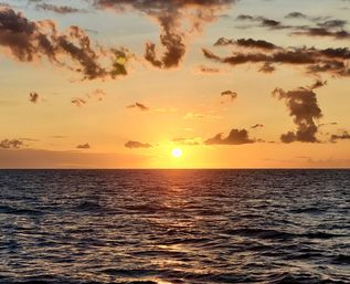 Golden sunset over the open ocean horizon, sun low above rippled water with shimmering reflections and scattered clouds.
