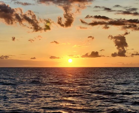 Golden sunset over the open ocean horizon, sun low above rippled water with shimmering reflections and scattered clouds.