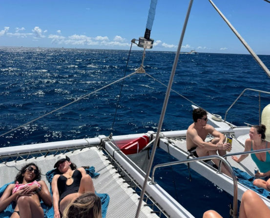 Group of people sunbathing and chatting on the trampoline nets of a catamaran sailboat on deep blue open ocean under a sunny sky.