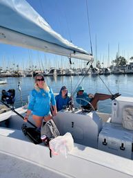 Three people in bright blue shirts relaxing on a sailboat at a sunny coastal marina — one smiling with sunglasses, one at the helm, and one lounging with legs up; calm water, clear blue sky, and rows of moored sailboats and masts in the background.