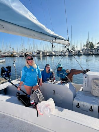Three people in bright blue shirts relaxing on a sailboat at a sunny coastal marina — one smiling with sunglasses, one at the helm, and one lounging with legs up; calm water, clear blue sky, and rows of moored sailboats and masts in the background.